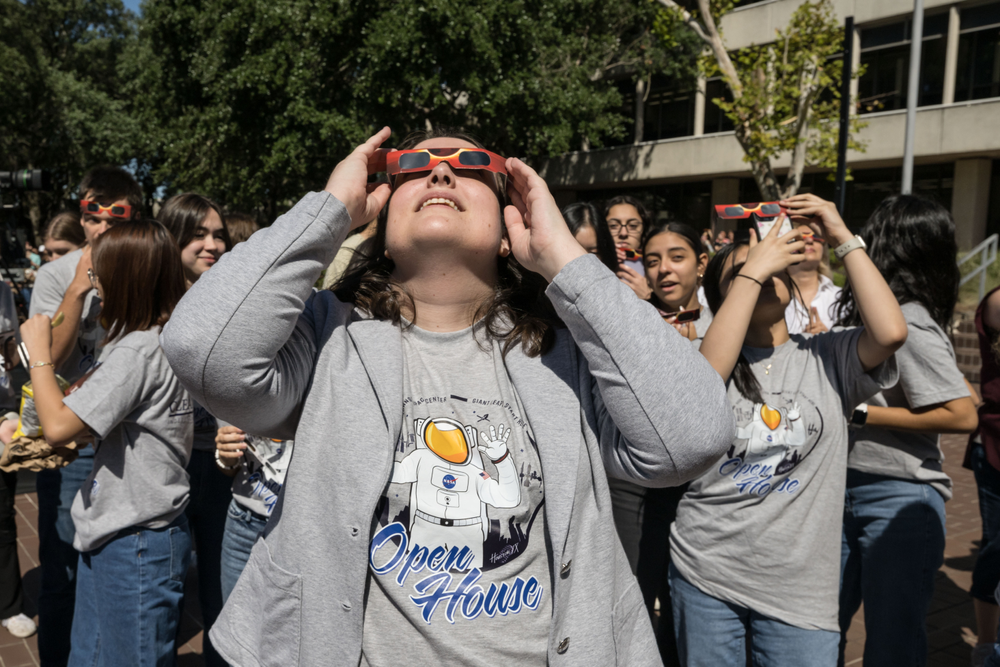 A person wearing eclipse glasses looking up at the sky in a crowd. She is wearing a grey shirt that has an astronaut and Open House logo on it with a grey sweater. 